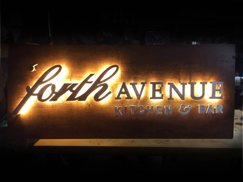an illuminated sign about 4 and a half feet wide by 2 feet high that reads 'forth avenue kitchen and bar' made from wood